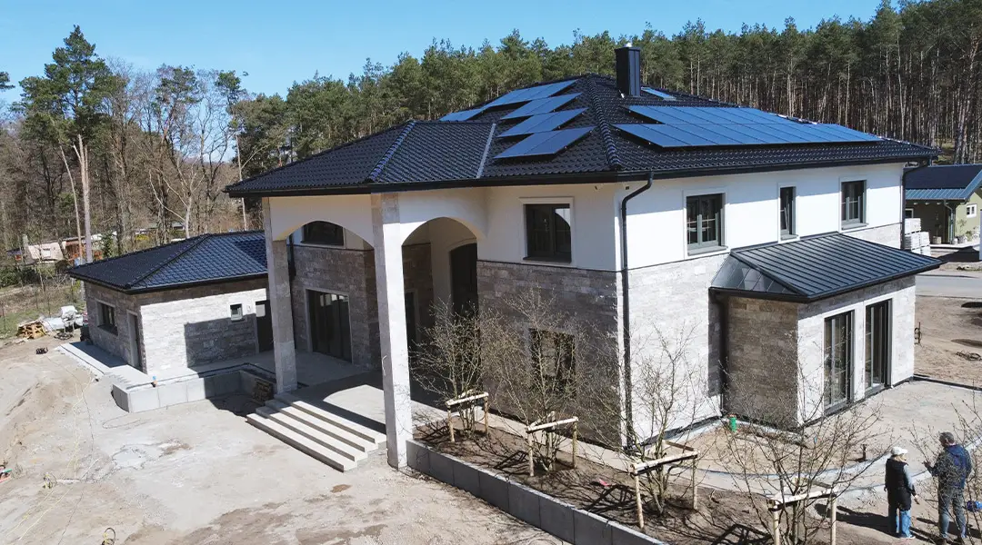 einfamilienhaus-neubau-photovoltaik-kirk-bau-01 Drohnenaufnahme eines modernen Wohnhauses mit Natursteinfassade, schwarzem Dach und Photovoltaikanlage.