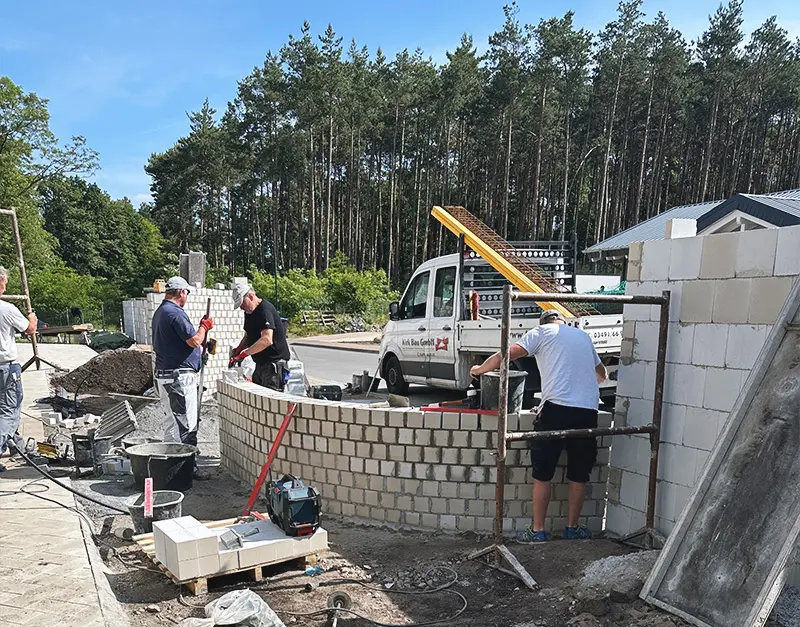 mauerarbeiten-rohbau-kirk-bau-01 Baustellenteam bei Maurerarbeiten zwischen Baumaterialien und vorbereiteten Wandflächen.
