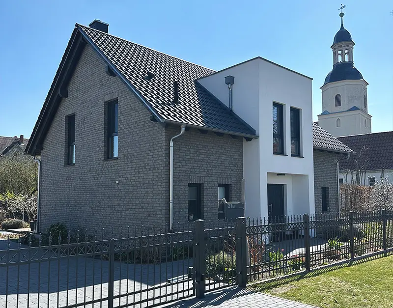 modernes-einfamilienhaus-klinkerfassade-kirk-bau-01 Modernes Einfamilienhaus mit grauer Klinkerfassade, weißem Anbau und Kirchturm im Hintergrund.
