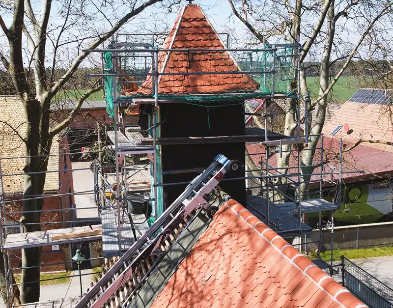 sanierung-historischer-gebaeudeturm-dacharbeiten-kirk-bau-01 Nahaufnahme eines eingerüsteten Gebäudeturms mit erneuerter roter Dacheindeckung.